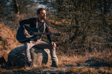 Man with an acoustic guitar is sitting on a stone in the nature