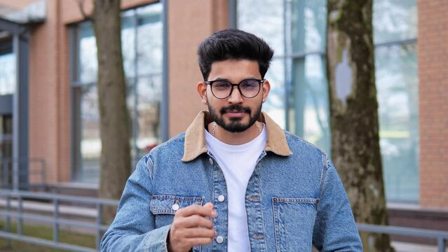 Happy trendy young cool man of Indian Hindu ethnicity is shown smiling to the camera while putting on his glasses. He is wearing a denim blue jacket suggesting a sense of adventure and freedom.