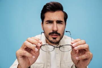 Close-up portrait of a brunette man looking through glasses that he holds in his hands, eye problems, glasses for vision farsightedness and nearsightedness, on a blue background, copy space 