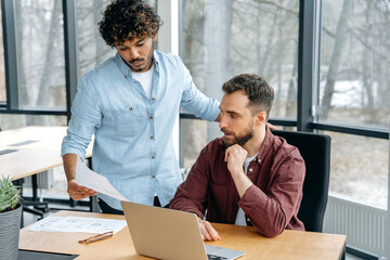 Two men of indian and caucasian ethnicity, work colleagues, work together on a project in a modern office, advise each other, share ideas and solutions, plan a product promotion strategy