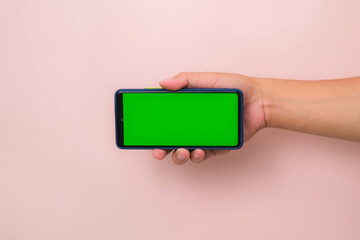 Human hand holding mobile smartphone with green screen in horizontal position isolated on pink background.