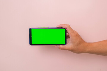 Human hand holding mobile smartphone with green screen in horizontal position isolated on pink background.