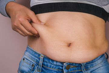 Naklejka premium Fat woman hand holding excessive belly fat isolated on pink background. Overweight fatty belly of woman. Female diet and body health care concept