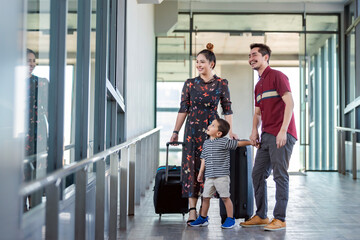 Happy young asian family travel of three walking with suitcases at airport terminal, Smiling parents with little son holding hands look out the window, Enjoying travelling together, Copy Space