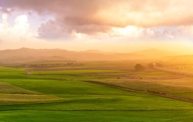 Obraz premium beautiful green valley with green fields with green spring grass with nive hills and mountains and scrnic colorful cloudy sunset on background of landscape