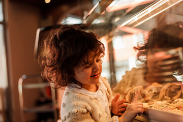 Cute little curly girl 3 years old with a young red-haired mother in the bakery at the counter chooses pastries. Cafe.