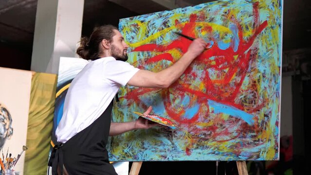 Contemporary artist with specialization in abstract art using brushes to create vibrant emotional oil painting on a large canvas Artist is displaying innovative talented use of colorful paint in work.