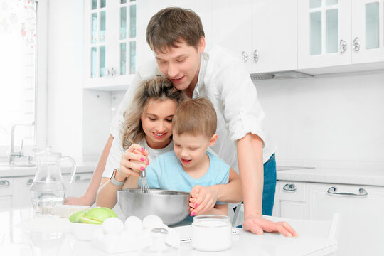 Young Caucasian Family Of Three Cooks Lunch Or Festive Cake In Kitchen According To Recipe. Happy Family At Table In Bright Kitchen..