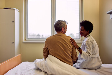 Obraz premium A female African-American doctor sitting with a male patient in a hospital, holding a hand on his shoulder.
