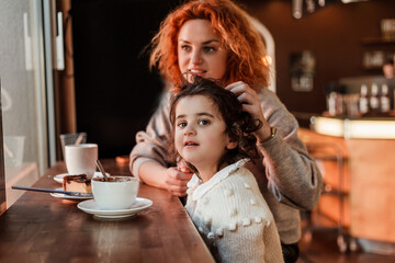 Beautiful young red-haired mother with cute curly-haired daughter are sitting in cozy cafe and drinking hot school. Mothers Day. Warm.