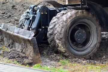 Fototapeta na wymiar Wheeled tractor with shovel clears ground on cloudy rainy day. Dirty bulldozer shovel close-up..
