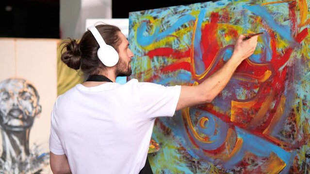 Creative artist wearing a white t-shirt black apron, with headphones on, is creating an abstract painting by painting it with vibrant colors while listening to favorite music and dancing in studio.