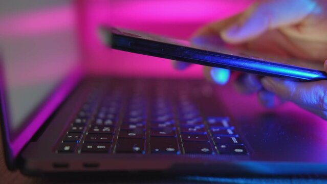 Use of mobile phone in trendy neon lights with laptop. Creative vivid color of ultraviolet red and blue. Hands of Girl scrolling up photos Close-up at dark room. App for sale in shop, Social media.