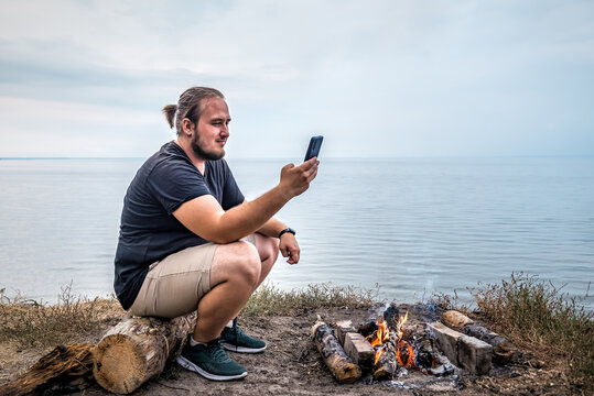 Young adult man sitting outdoors lakeshore near campfire with his smartphone