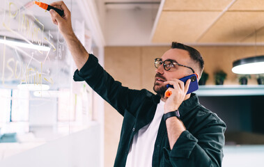 Serious man with smartphone and highlighter writing on glass board