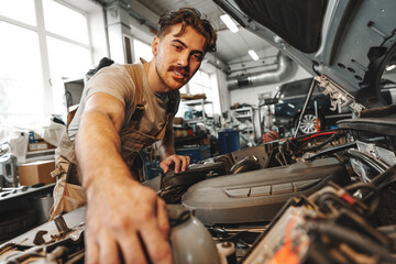 Young male mechanic examining engine under hood of car at the repair garage - obrazy, fototapety, plakaty