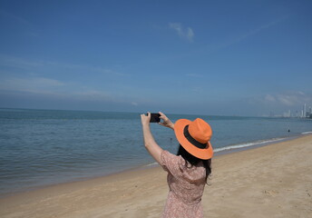 A adult female tourist Use both hands to lift up your mobile phone to capture the beauty of sea on a sunny day. A woman wearing light brown dress, an orange wide-brimmed hat is happy to take pictures.