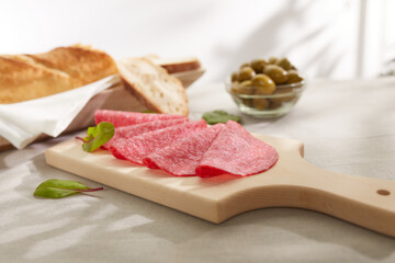 salami fine slices with bread and olives. sausage and herbs on a wooden board. snack on a light table. meat delicacy under sunlight. dried sausage serving. 