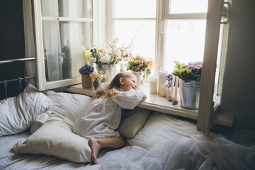 Girl in white dress sitting on a bed near window with lots of flowers.