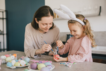 Obraz na płótnie Canvas Happy mother and her little daugter decorating Easter eggs using brush and paint on the kitchen.