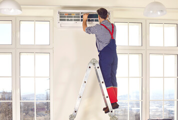 Professional electrician man standing back on a ladder maintaining or cleaning modern air conditioner indoors. Mature technician in the room repairing or installing air conditioner.