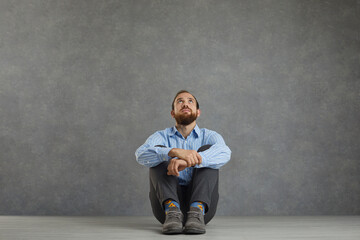 Relaxed adult pensive thoughtful caucasian man with praying face sit on floor looking up and dreaming about future waiting for miracle studio shot. Male full-length portrait. Thoughtfulness, wonder
