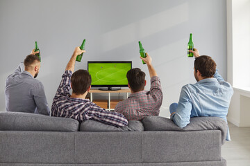 Group of men with beer watching live football game on TV. 4 young friends sitting on comfortable sofa, holding green beer bottles and watching soccer championship on television, backside rear view
