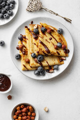 Crepes or blinis or thin pancakes with blueberries, hazelnuts and chocolate sauce on a white plate at white stone table. Maslenitsa traditional festival meal.