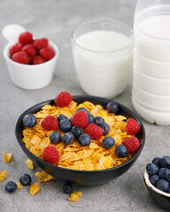 Tasty corn flakes with blueberries in a black bowl with milk on grey background, close up.