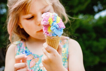 Happy preschool girl eating colorful ice cream in waffle cone on sunny summer day. Little toddler...