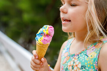 Happy preschool girl eating colorful ice cream in waffle cone on sunny summer day. Little toddler...