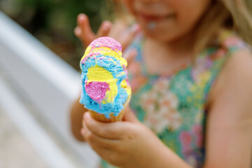 Happy preschool girl eating colorful ice cream in waffle cone on sunny summer day. Little toddler...