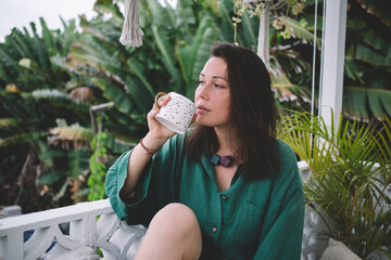 Contemplative hipster girl 30sl holding tea cup recreating during coffee break at balcony, pensive...