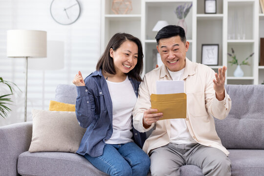 Cheerful and happy asian family together, man and woman at home on sofa sitting in living room, received postal envelope with good news, message and good achievement news. - Powered by Adobe