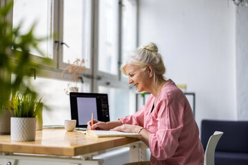 Smiling mature businesswoman writing in notebook while sitting at table in office
