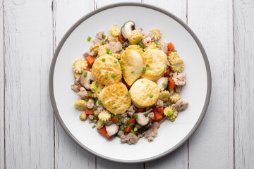 Stir fried egg tofu and chicken in oyster sauce with corn, carrot and shiitake in white dish on wooden table.