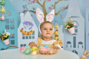 Obraz na płótnie Canvas Baby girl celebrate Easter. Funny happy kid in bunny ears Sits by the table and playing with Easter eggs. Colorful Easter eggs and flowers. Home decoration and flowers