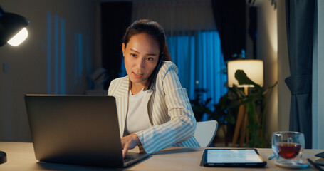Fototapeta na wymiar Young happy Asian businesswoman wear formal sit in front of desk with laptop and tablet typing and call smartphone meeting with colleague in cozy living room at home night. Overtime work concept.