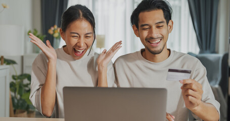 Happy young Asian couple sit on couch with laptop success credit card payment online shopping at home on holiday. Smile young husband and wife excited browsing app on gadgets. Lifestyle concept.