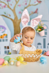 Baby girl celebrate Easter. Funny happy kid in bunny ears Sits by the table and playing with Easter eggs. Colorful Easter eggs and flowers. Home decoration and flowers