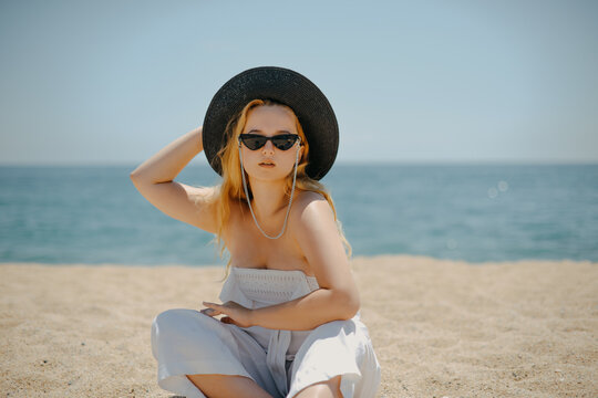 Fashion photo of a girl in black glasses with a chain and a large straw hat, against the backdrop of the sea or ocean. Woman sunbathes on the beach, summer photo. Blonde with long hair