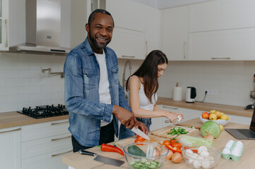 A happy couple preparing food in the kitchen. the African-American man looks on, smiling. the couple prepares a salad