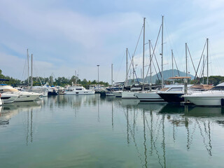 Luxury boats in marina. Yacht club. Sailboats on the harbour. Reflection of masts on a sea surface. Calm bay. Sky with clouds. Moored vessels. Tropical islands. Land on the horizon. Sailing, yachting