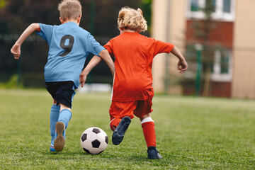 Obraz premium Two school boys kicking a soccer ball. School boys in red and blue football uniforms compete in a soccer match. Football duel in children soccer game