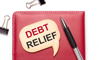 On a light background there are black paper clips, a pen, a burgundy notepad a wooden board with the text DEBT RELIEF