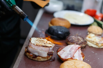 Cook using burner flame to prepared burger in a restaurant kitchen. American diner chef preparing hamburgers with melted cheese and bacon