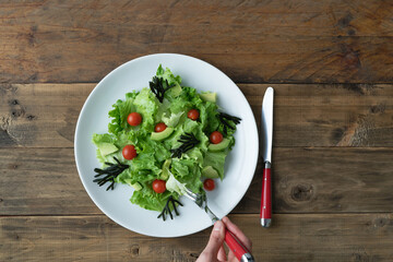 Hand with fork eating assorted salad on white plate on wooden background. Copy space.