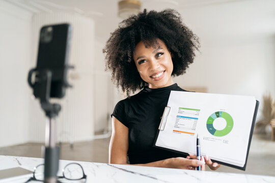 A cheerful young curly-haired woman in the office stream online using the phone shows financial charts. - Powered by Adobe