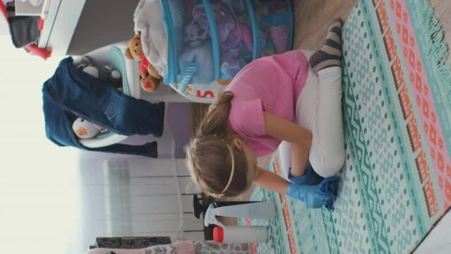 Vertical video. Little girl in rubber gloves doing housework, cleaning the rug in her messy room