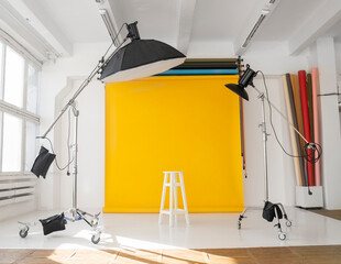 Yellow background roll with a white chair and two professional flash on a c-stands. Sunny daylight photo studio interior with set of professional equipment.